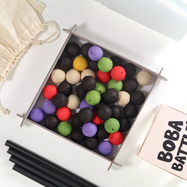 Top-down view of the Boba Battle by Pikkii party game setup, featuring a game arena filled with colorful cotton paper boba balls, black straws, a drawstring bag, and the game box.