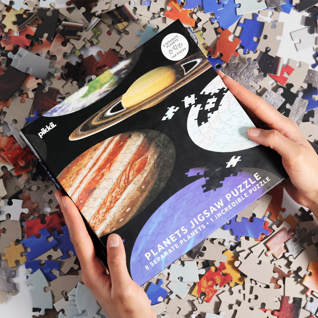 Hands holding planets jigsaw puzzle packaging by Pikkii on top of puzzle pieces scattered